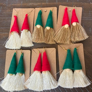 May include: Set of six pairs of gnome-themed tassel earrings. Each pair features a felt gnome hat in either red or green with a wooden bead face and a white tassel.