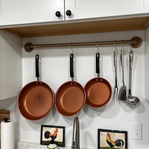 Puede incluir: Un estante de utensilios de cocina de metal dorado con ganchos que cuelgan de una tubería. Cuatro sartenes de color cobre y tres utensilios de plata cuelgan de los ganchos. El estante está montado en una pared blanca sobre una encimera blanca con un fregadero de acero inoxidable. Dos azulejos decorativos con temática de gallo están en la pared debajo del estante.