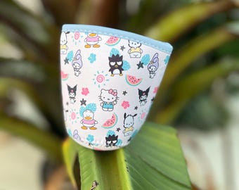 May include: A white cup cozy with a light blue trim, decorated with cartoon characters, watermelon slices, and floral accents. The cozy is designed to fit around a cup or mug.