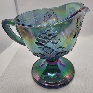 May include: Iridescent blue glass creamer with a pedestal base. The creamer features a grape and leaf design, with a handle and a pouring spout. The glass has a shimmering, multi-colored finish.