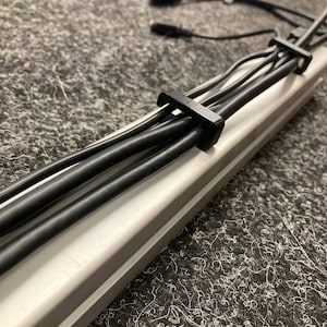 以下が含まれることがあります： ワイヤーを整理するための黒いケーブルクリップ。クリップは金属製のチャンネルに取り付けられており、複数の黒いケーブルを固定しています。