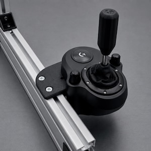May include: A black gear shifter with a cylindrical handle and a leather-like boot, mounted onto a silver aluminium extrusion profile. The shifter features a circular base with a prominent logo button and two small knobs, secured by metal screws.