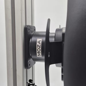 Puede incluir: Soporte vertical plateado con un soporte de volante de carreras MOZA negro fijado. El soporte está asegurado con tornillos y presenta el logotipo de MOZA. La imagen también incluye el texto "BALTIC MOTORSPORT SOLUTIONS" y un gráfico de un piloto de carreras.