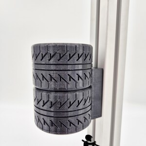 May include: Black 3D-printed tyre holder with a mounting bracket, designed to hold two tyres. The holder is attached to a silver metal support. The text "BALTIC MOTORSPORT SOLUTIONS" is at the bottom.