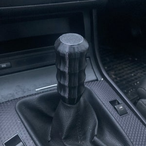 Peut inclure: Un pommeau de levier de vitesses noir et texturé avec un design cylindrique, installé dans une voiture. Le pommeau a une prise rainurée et un dessus plat. L'image montre également l'intérieur de la voiture, y compris le soufflet de levier de vitesses et le tableau de bord.