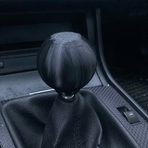 May include: Black spherical gear shift knob with a textured surface. The knob is attached to a black leather gear shift boot. The text "BALTIC MOTORSPORT SOLUTIONS" is visible at the bottom of the image.
