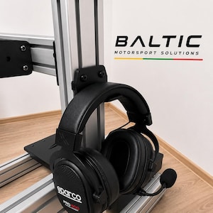 Pode incluir: Auscultadores de corrida pretos Sparco Pro 2000 com microfone, montados num suporte de perfil de alumínio prateado. O texto BALTIC MOTORSPORT SOLUTIONS é visível a preto com uma linha amarela, verde e vermelha por baixo na parede branca.