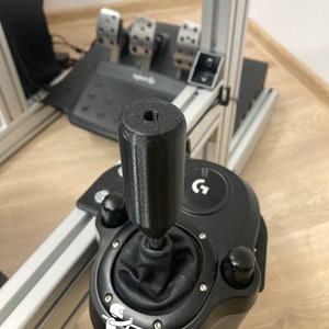 Könnte beinhalten: Schwarzer Schalthebel mit einem Leder-Schaltknauf und einem schwarzen Sockel mit einem silbernen G-Logo. Der Schalthebel hat einen schwarzen Knauf mit einem kleinen Loch oben.