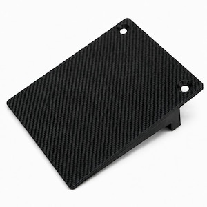 May include: A rectangular black carbon fiber panel with a woven texture, featuring two countersunk mounting holes along one edge and a perpendicular support bracket on the underside. The text BALTIC MOTORSPORT SOLUTIONS is printed below.