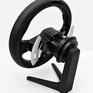 Può includere: Un volante da corsa nero con impugnatura traforata, leve del cambio argentate e logo Fanatec centrale, montato su un supporto nero con trama in fibra di carbonio. Il testo BALTIC MOTORSPORT SOLUTIONS è presente alla base dell'immagine.