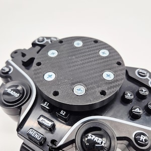 Może przedstawiać: Zbliżenie czarnego kontrolera kierownicy wyścigowej z wieloma przyciskami i środkiem z włókna węglowego. Przyciski to m.in. "Radio", "Menu" i "Start". Tekst "Baltic Motorsport Solutions" znajduje się na dole.