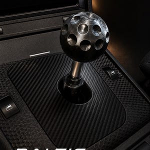 May include: A black spherical gear shift knob with a dimpled surface texture, mounted on a metallic shaft. The knob is positioned on a black carbon fiber center console panel featuring window switches and the text BALTIC MOTORSPORT SOLUTIONS.