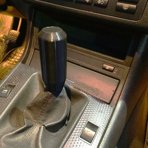 May include: Black gear shift knob with a textured surface. The knob is installed in a car's gear shift lever. The gear shift lever is surrounded by a black leather boot. The car's interior is visible in the background.