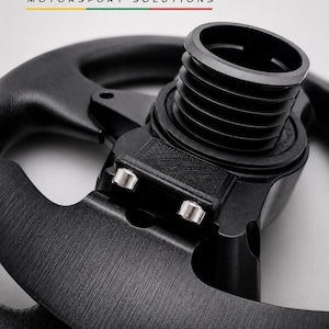 May include: A black racing steering wheel hub adapter featuring a central cylindrical mount with ribbed sides and two silver metallic pins. The text BALTIC MOTORSPORT SOLUTIONS is printed above the component in black, red, and green.