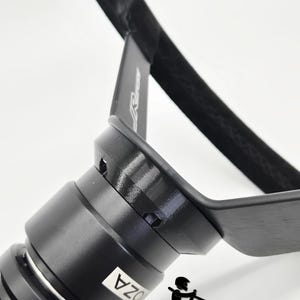 May include: Close-up of a black steering wheel with a textured grip. The wheel has a cylindrical base with the text "OZA" on a white label. The words "BALTIC MOTORSPORT SOLUTIONS" are printed at the bottom, along with a small graphic of a race car driver.