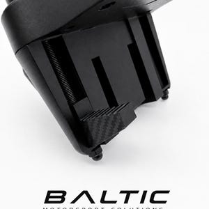 Può includere: Un pezzo di ricambio per morsetto di fissaggio del cambio in nero opaco con un'asta di regolazione filettata e una superficie di presa con motivo in fibra di carbonio. Il componente riporta il testo BALTIC MOTORSPORT SOLUTIONS.