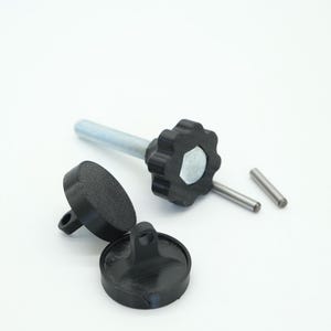 Pu&ograve; includere: Una collezione di componenti hardware neri, tra cui un bullone filettato con una manopola a forma di stella, due piccoli perni cilindrici e due pezzi circolari con un anello. I componenti sono su sfondo bianco.