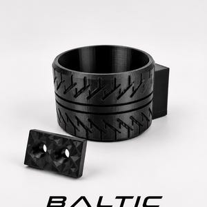 May include: A black cylindrical cup holder with a textured tire tread pattern on the exterior and a separate rectangular mounting plate featuring two countersunk screw holes. The text BALTIC MOTORSPORT SOLUTIONS is printed below.