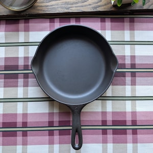May include: A dark grey cast iron skillet with a long handle and a rounded cooking surface. The skillet is set on a checkered tablecloth with red, white, and green stripes. The number "6" is visible on the handle.