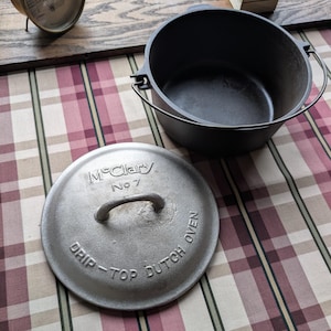 May include: A black cast iron Dutch oven with a matching lid. The lid features the embossed text McClary No 7 and DRIP-TOP DUTCH OVEN, along with a central handle. The pot includes a wire bail handle attached to the sides for lifting and carrying.