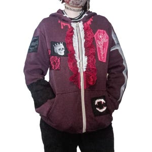 May include: A burgundy zip-up hoodie with patches, including a skull and coffin design. The hoodie features a ruffled trim and black accents on the sleeves and pockets. A face mask with a blood splatter design is also visible.
