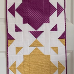 May include: A quilted wall hanging with a geometric design. The design features three squares with different patterned fabrics. The top square is green, the middle square is purple, and the bottom square is yellow. The squares are surrounded by white triangles.