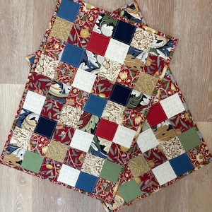 May include: Two square patchwork quilted placemats. Each placemat features a red floral patterned fabric as a base, with a grid of squares in various colors and patterns, including red, white, blue, and green. The placemats are designed for dining.