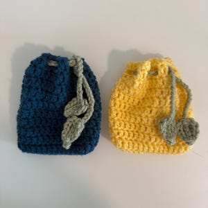 May include: Two handmade crochet drawstring pouches. One is navy blue, the other is yellow. Both have a matching green drawstring with leaf-shaped embellishments. The pouches are likely used for small items or gifts.