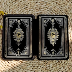 May include: A black leather-bound journal with gold accents and a decorative design featuring a bird in flight. The journal is closed and lying on a woven straw surface.