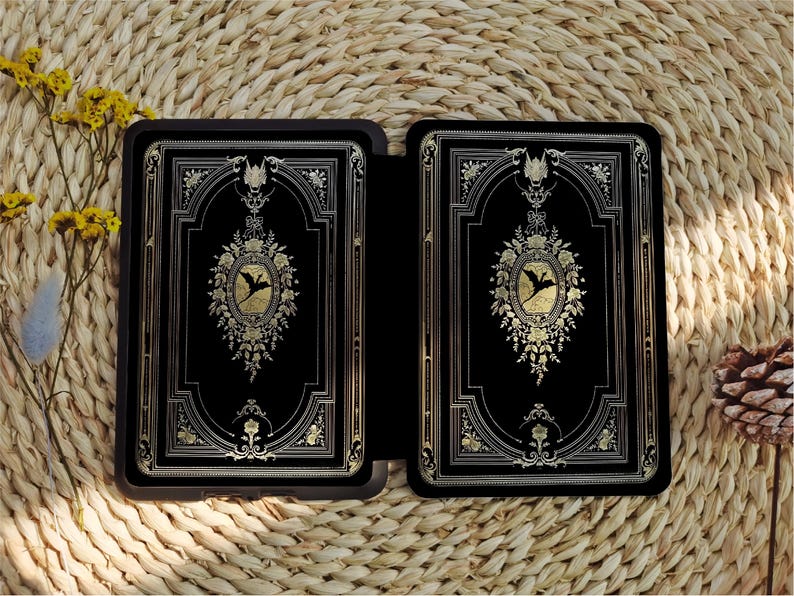 May include: A black leather-bound journal with gold accents and a decorative design featuring a bird in flight. The journal is closed and lying on a woven straw surface.