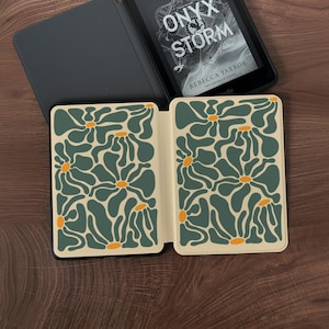 May include: An open Kindle e-reader with the book "Onyx Storm" by Rebecca Yarros displayed. The cover is in a black case with a floral design in green and orange on a cream background. The e-reader is on a wooden surface.