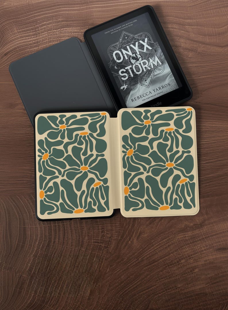 May include: An open Kindle e-reader with the book "Onyx Storm" by Rebecca Yarros displayed. The cover is in a black case with a floral design in green and orange on a cream background. The e-reader is on a wooden surface.