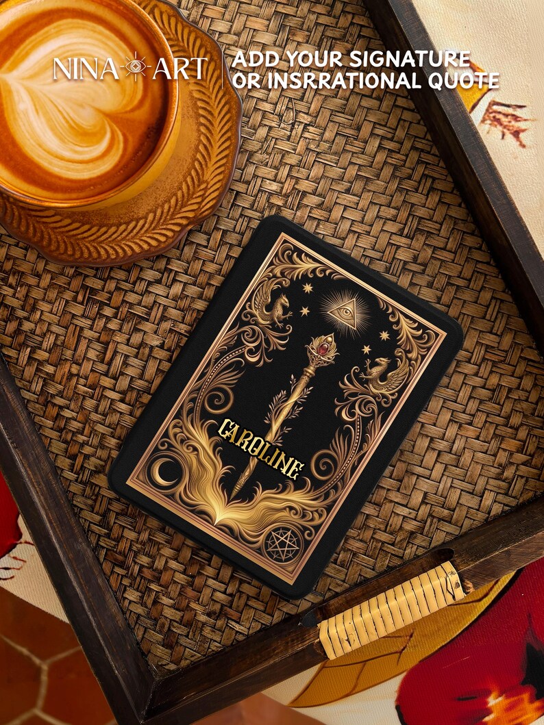 Op de afbeelding: Een zwart en goud decoratief item met de naam "CAROLINE", met sierlijke ontwerpen, een staf en hemelse symbolen. Het item ligt op een geweven oppervlak, met een kop koffie op de achtergrond. De tekst "NINA ART" en "ADD YOUR SIGNATURE OR INSRRATIONAL QUOTE" zijn ook zichtbaar.