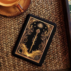 Op de afbeelding: Een zwarte tarotkaart met gouden sierlijke ontwerpen, waaronder een staf, sterren en een oog in een driehoek. De kaart ligt op een geweven dienblad naast een kopje koffie met latte art.