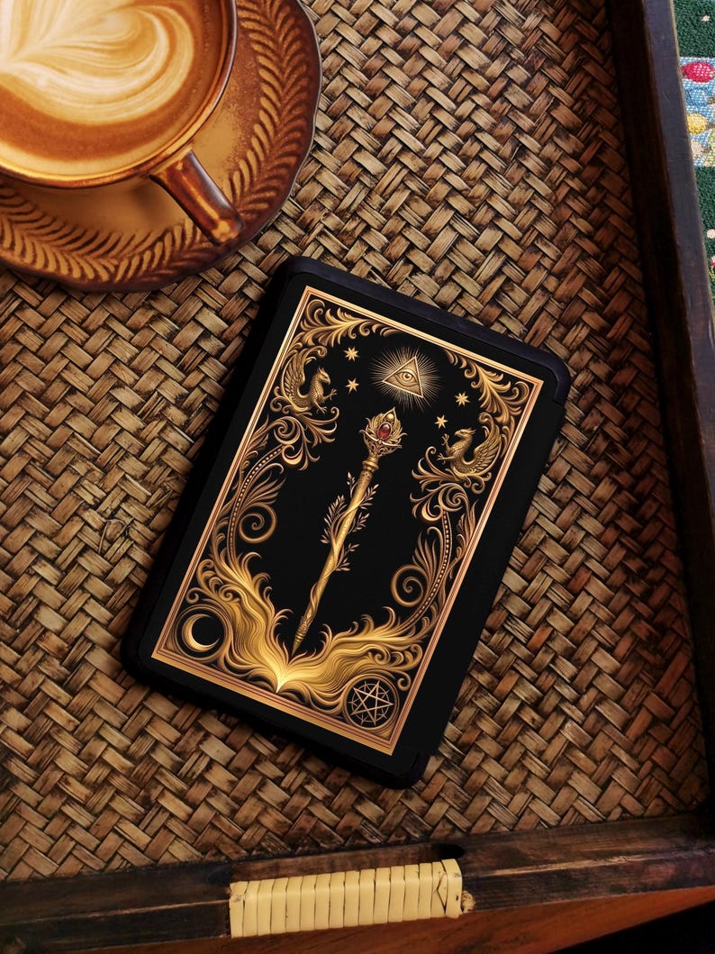 Op de afbeelding: Een zwarte tarotkaart met gouden sierlijke ontwerpen, waaronder een staf, sterren en een oog in een driehoek. De kaart ligt op een geweven dienblad naast een kopje koffie met latte art.