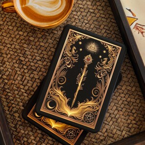 Op de afbeelding: Twee zwarte tarotkaartendecks met gouden sierlijke ontwerpen met een staf, sterren en een oogsymbool. Een kopje koffie met latte art staat in de buurt op een geweven dienblad. De kaarten hebben een maan- en sterrenpatroon.