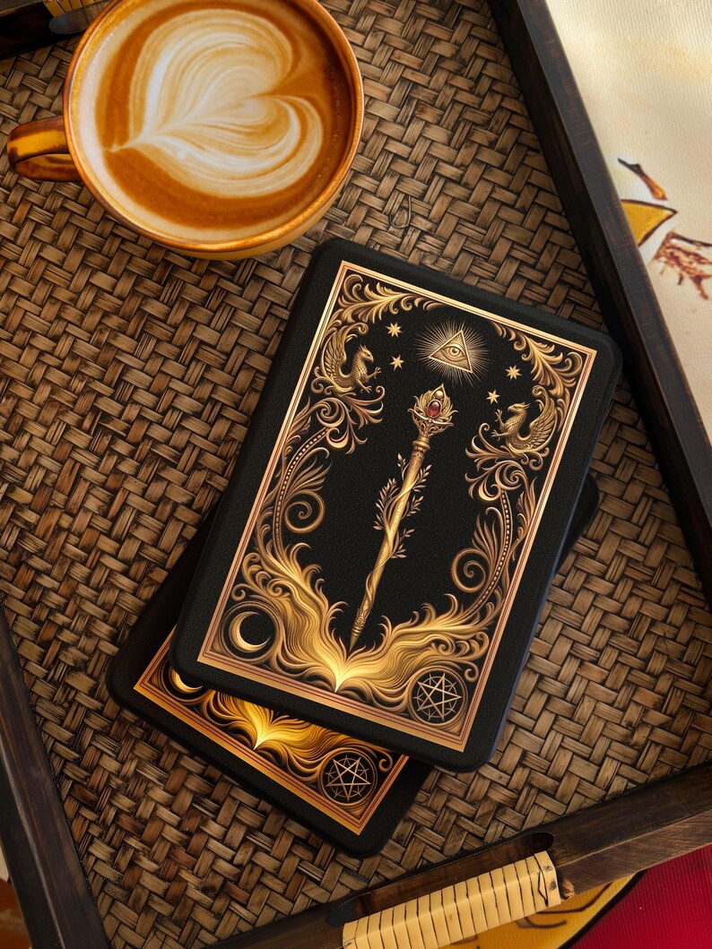 Op de afbeelding: Twee zwarte tarotkaartendecks met gouden sierlijke ontwerpen met een staf, sterren en een oogsymbool. Een kopje koffie met latte art staat in de buurt op een geweven dienblad. De kaarten hebben een maan- en sterrenpatroon.