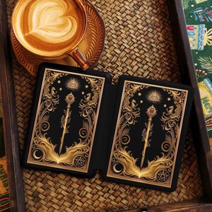 Op de afbeelding: Een kop koffie met latte art staat op een schotel naast een zwarte tarotkaarthouder met gouden sierlijke ontwerpen. De kaarten hebben een gouden staf met een rode edelsteen, sterren en een symbool.