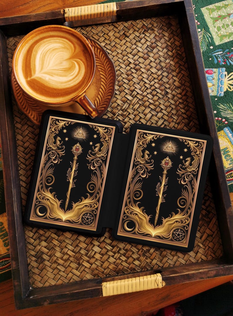 Op de afbeelding: Een kop koffie met latte art staat op een schotel naast een zwarte tarotkaarthouder met gouden sierlijke ontwerpen. De kaarten hebben een gouden staf met een rode edelsteen, sterren en een symbool.