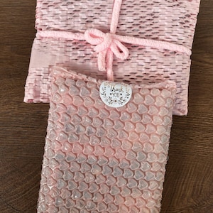Op de afbeelding: Roze verpakkingsmaterialen, waaronder bubbeltjesplastic en geweven papier, zijn gerangschikt op een houten oppervlak. Een roze koord is om het geweven papier gebonden en vormt een strik. Een sticker met de woorden "Thank You" is zichtbaar.