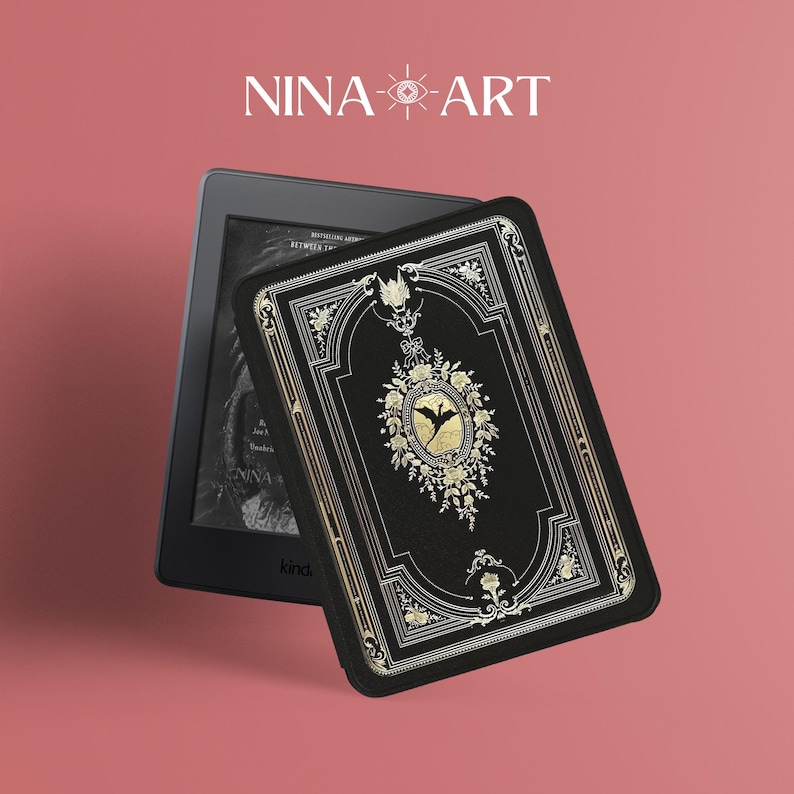 May include: A black Kindle e-reader with a black and gold decorative cover featuring an ornate design with a bat and floral accents. The cover has the text "NINA-ART" on the front.