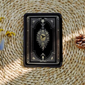 May include: A black leather-bound journal with gold accents and a decorative design featuring a bird in a floral wreath. The journal is closed and lying on a woven straw surface.