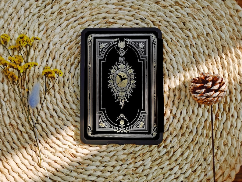 May include: A black leather-bound journal with gold accents and a decorative design featuring a bird in a floral wreath. The journal is closed and lying on a woven straw surface.