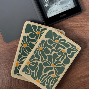 May include: Two rectangular coasters with a floral pattern in shades of green and orange on a beige background. A Kindle e-reader with a book cover visible is in the background. The book cover reads "ON A STORM" by Rebecca Yarros.