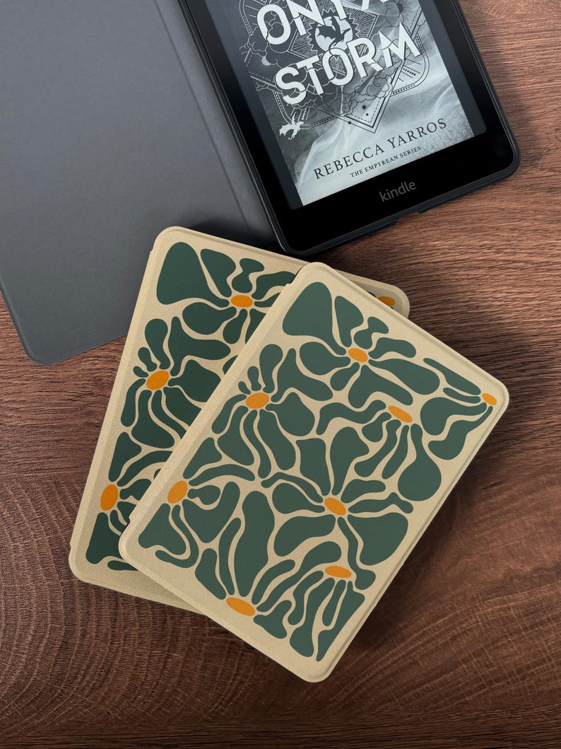 May include: Two rectangular coasters with a floral pattern in shades of green and orange on a beige background. A Kindle e-reader with a book cover visible is in the background. The book cover reads "ON A STORM" by Rebecca Yarros.