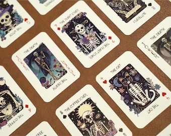 Puede incluir: Una baraja de cartas con un tema gótico y espeluznante. Cada carta presenta un esqueleto diferente con un diseño único. Las cartas están impresas en cartulina blanca con tinta negra. Las cartas están dispuestas en forma de abanico sobre una superficie marrón.