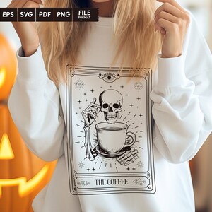 May include: A white sweatshirt with a black and white illustration of a skeleton holding a coffee cup. The illustration is in a vintage style frame with the text "The Coffee" at the bottom.
