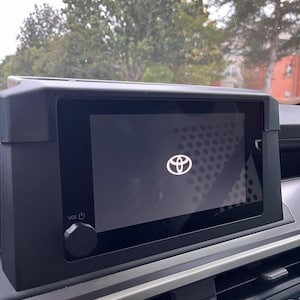 May include: A black car infotainment system with a screen displaying the Toyota logo. The system has a volume knob and is set in a car's dashboard. The background shows trees and a building.