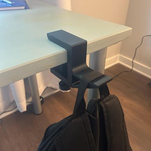 Peut inclure: Un serre-joint de bureau noir avec un sac à dos suspendu. La pince est fixée au bord d'un bureau bleu clair. Le sac à dos est noir et possède des bretelles réglables.