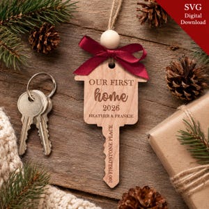 May include: A wooden key-shaped ornament featuring a dark red ribbon and a round wooden bead. The ornament is engraved with the text "OUR FIRST home 2026", "HEATHER & FRANKIE", and "100 FIELDSTONE PLACE". A metal keyring with two keys is attached.